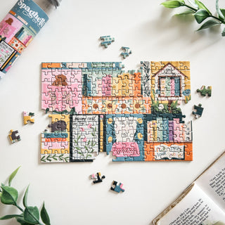 The One More Chapter 150 Piece Jigsaw Puzzle by Spaghetti Tree features illustrated text and drawings on a white surface, with scattered pieces and green plant leaves nearby.