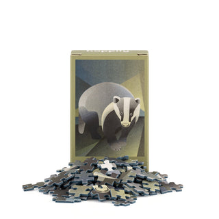 A box featuring an illustration of a badger sits behind a scatter of jigsaw puzzle pieces against a white background, highlighting the Badger | 99 Piece Mini Jigsaw Puzzle by Happily Jigsaw Puzzles—a delightful mini puzzle and an ideal wildlife gift for nature lovers.