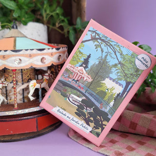 A pink box labeled Balade Au Jardin Public | 100 Piece Jigsaw Puzzle by Pièce Rapportée, featuring a park scene, leans against a carousel with a checkered cloth partially visible in front.