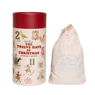 A cylindrical container labeled "123 Farm Christmas | 500 Piece Jigsaw Puzzle" sits beside a drawstring cloth bag featuring blue text and a subtle graphic.