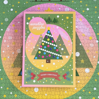 The Sunshine Puzzles Merry Christmas | 1,000 Piece Jigsaw Puzzle features a limited edition rectangular box with a decorated Christmas tree, displayed atop the completed festive puzzle.