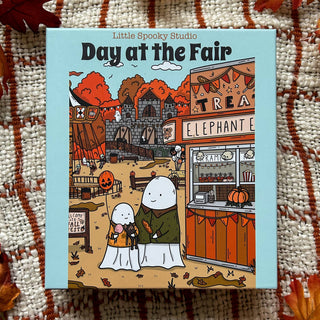 The illustrated game box for Little Spooky Studio’s Day At the Fair | 1,000 Piece Jigsaw Puzzle features two ghost characters enjoying a fall fair surrounded by food stands, pumpkins, and autumn trees, creating a charmingly spooky autumn scene.