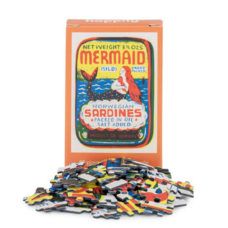 A box with Louise Lockhart’s retro sardines design labeled “Mermaid | 99 Piece Mini Jigsaw Puzzle” from Happily Jigsaw Puzzles stands behind a pile of loose puzzle pieces from this charming mini puzzle.