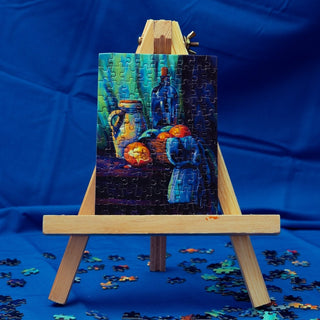 The Minizz Still Life | 99 Piece Jigsaw Puzzle, featuring bottles, jars, and fruit, stands finished on a small wooden easel with loose pieces nearby, all set against a blue cloth background.
