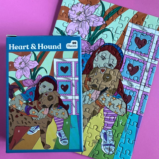 A partially assembled matte finish Heart & Hound | 99 Piece Jigsaw Puzzle and its box, featuring an illustration of a person hugging a spotted dog amidst flowers and heart shapes.