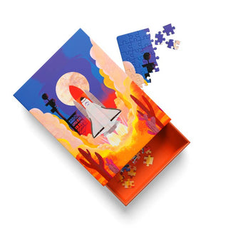 The “Space Shuttle | 99 Piece Jigsaw Puzzle” by Ohh Deer features a partially assembled rocket launch scene, radiating cosmic energy as it rests beside its vibrant orange box filled with more pieces—evoking the thrill of a NASA mission underway.