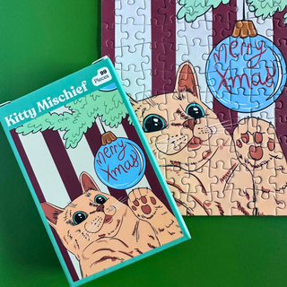 A matte finish pocket-sized Kitty Mischief | 99 Piece Jigsaw Puzzle box next to a partially assembled puzzle featuring a cat reaching for a Merry Xmas ornament on a striped background.