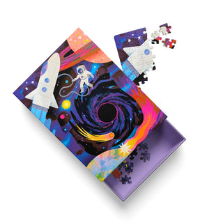 The Astronaut | 99 Piece Jigsaw Puzzle by Ohh Deer features a vibrant space theme with a rocket, astronaut, and black hole. It creates cosmic vibes as it sits partially assembled, with several pieces scattered in and around its distinctive purple box.
