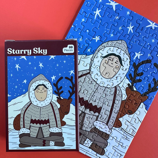 The Starry Sky | 99 Piece Jigsaw Puzzle box sits next to the completed puzzle showing a cartoon person in a parka and a reindeer under a starry night sky with a matte finish.