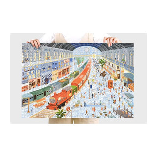 A person holds the completed Time Station | 1,000 Piece Jigsaw Puzzle by Momibook (19.7" x 29.5"), featuring a lively train station with multiple trains, people, shops, and an arched glass roof.