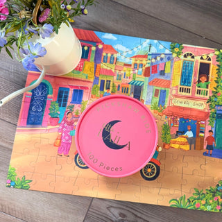 A pink "Rickshaw Ride | 100 Piece Jigsaw Puzzle" by Laila Luxury Puzzles rests on the completed Asma Jamali market street puzzle, capturing a vibrant Pakistan scene, beside a white flower jug.