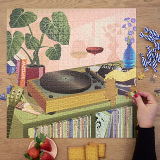A hand adds a piece to the completed ViSSEVASSE "Music & Me" 500-piece jigsaw puzzle, showing a turntable, books, plant, flower vase, and snacks on a wooden table.
