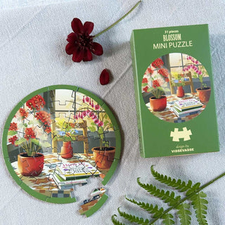 A vibrant, round 31-piece jigsaw puzzle from ViSSEVASSE, featuring a lively plant scene, rests beside its “Blossom | 31 Piece Jigsaw Puzzle” box on a white surface, accompanied by a red flower and fern leaf—making this compact travel puzzle ideal for puzzlers on the move.