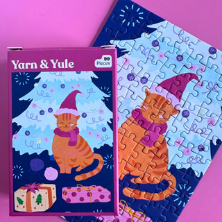 The Yarn & Yule | 99 Piece Jigsaw Puzzle box shown with its completed mini puzzle of an orange cat wearing a winter hat and scarf in front of a Christmas tree and gifts.