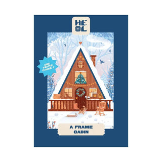 Illustration of an A-frame cabin in snow with a wreath, snowman, rocking chair, and trees. Text: "A-Frame Cabin." Heol Editions' A-Frame Cabin | 99 Piece Jigsaw Puzzle is perfect for Christmas and made in France.