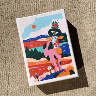 A colorful illustration of a cow in a vibrant landscape with mountains, flowers, and a blue sky—designed by Katharina Bourjau—adorns the rectangular box of the Almsommer | 99 Piece Jigsaw Puzzle from Das Puzzle Kollektiv, printed on recycled paper and shown resting on a textured beige surface.