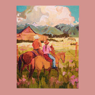 Two riders on horseback face a barn and distant mountains amid green fields and wildflowers under a partly cloudy sky in Saddle Up, a stunning 500 piece western landscape jigsaw puzzle from WerkShoppe.
