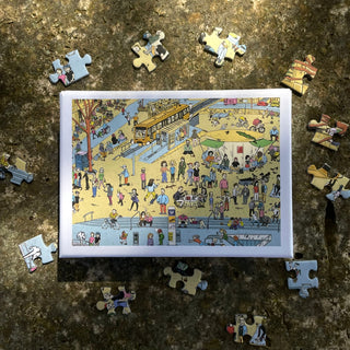 A rectangular recycled paper box from Das Puzzle Kollektiv holds the Marienplatz 2.0 | 99 Piece Jigsaw Puzzle, featuring Nils Knoblich’s vibrant illustration; it rests on a textured surface with several puzzle pieces scattered around it.