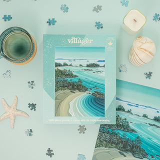 The Soaring Over Tofino 1,000-piece jigsaw puzzle by Villager Puzzles, featuring ocean and forest art by Dana Statham, rests on a mint surface with scattered pieces, seashells, and starfish—a coastal scene brought to life.