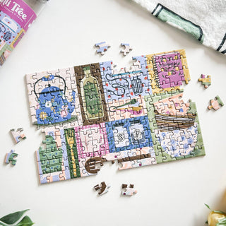 The partially completed From Scratch | 150 Piece Jigsaw Puzzle by Spaghetti Tree lies on a white surface, showcasing illustrated objects and labels, with several loose pieces scattered nearby.