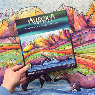 A hand holds an Aurora Puzzles "True Love" 1,000-piece eco-friendly jigsaw puzzle box with moose and colorful mountains; the finished puzzle with the same image is displayed in the background.