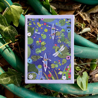 A boxed 99-piece jigsaw puzzle from Das Puzzle Kollektiv, titled “Canopy Canoeing,” features an illustration of kayakers, lily pads, and dragonflies. Resting on green garden hoses among leaves, it’s the perfect gift for outdoor enthusiasts and fans of Canopy Canoeing.