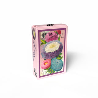 The Penny Puzzle "Asters Seeds" is a pink rectangular box with a hand-drawn illustration of colorful asters on the front, containing a charming 99-piece mini jigsaw puzzle inspired by vintage seed bags.