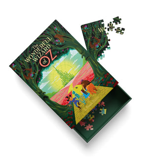 A 99-piece jigsaw puzzle from Ohh Deer, titled The Wonderful Wizard of Oz, featuring cover art by Andrew Kolb that depicts characters on the yellow brick road, with the partially assembled puzzle pieces scattered around the open box.