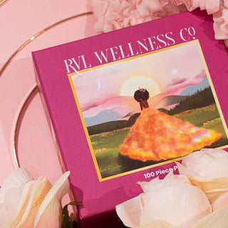 The Ophelia Aurelia 100 Piece Jigsaw Puzzle by RVL Wellness Co. features art of a woman in a glowing dress amid white flowers, perfect for self-care and relaxing puzzle time.