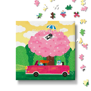 Blooming Drive | 196 Piece Jigsaw Puzzle by The Puzzle Theory features an illustration of a pink car, a person, and a cat beneath a blooming cherry tree, with scattered pieces around the box. Made from recycled paperboard, this charming springtime puzzle offers eco-friendly fun.