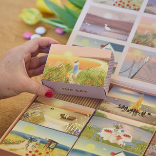 A hand holds a ViSSEVASSE Summer Moments Jigsaw Puzzle Box Set featuring a 104-piece puzzle with magnetic closure and an illustration of a person by a sunset lake; other puzzles from the set are arranged nearby.
