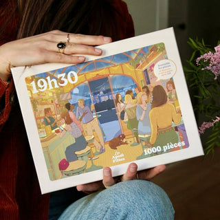 A person holds the Les Apero Filles 1,000 piece jigsaw puzzle by Lundi en Huit, featuring an illustrated scene of friends socializing in a lively cafe—an ideal choice for those who love friendship-themed puzzles.