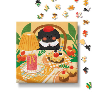 Enjoy assembling the Warm Wishes | 196 Piece Jigsaw Puzzle by The Puzzle Theory, featuring a charming illustration of a black dog in a red hat beside a picnic table filled with desserts, a drink, and a lamp—with scattered puzzle pieces floating above. Thoughtfully made from recycled paperboard for an eco-friendly birthday puzzle experience.
