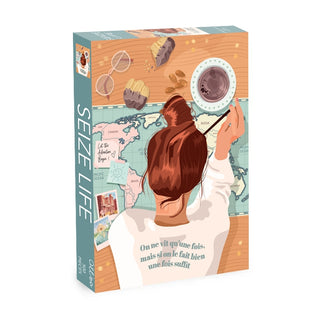 A woman with a bun writes on a world map at a table set with coffee, muffins, glasses, and postcards. The box reads “Seize Life,” and her shirt features a French quote. This charming scene is beautifully captured in the Seize Life | 500 Piece Jigsaw Puzzle by Olleo Puzzles, which also includes a cotton bag for convenient storage.
