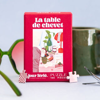 A red box labeled “La Table de Chevet | 100 Piece Jigsaw Puzzle” by Jour Férié—a delightful French-made bedside table puzzle—is displayed beside a green mug, green sunglasses, and two puzzle pieces on a light blue surface.