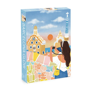 Barcelona Glow | 500 Piece Jigsaw Puzzle by Olleo Puzzles features an illustrated woman photographing colorful, mosaic-covered buildings inspired by Antoni Gaudí at sunset in Barcelona.