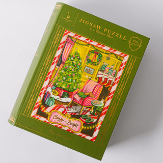 Box of a 1,000-piece jigsaw puzzle from Good Juju Ink, featuring a festive room with a decorated Christmas tree, gifts, cozy armchair, and mantle. Titled "Cozy & Bright," this holiday puzzle is perfect for bringing friends and family together during the season.