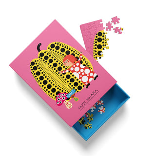 A pink box from Ohh Deer’s Yayoi Kusama 99 Piece Jigsaw Puzzle, decorated with a yellow and black polka-dotted pumpkin and a doll, sits nearby with colorful puzzle pieces partially assembled and scattered around.