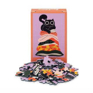 The Pillow Cat | 99 Piece Mini Jigsaw Puzzle by Happily Jigsaw Puzzles features a black cat atop colorful objects and comes in a box with a charming cat illustration. It's made from recycled board.