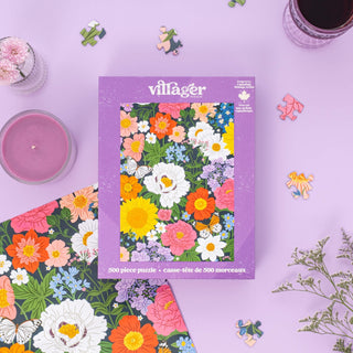The Midnight Blooms | 500 Piece Jigsaw Puzzle by Villager Puzzles features a vibrant floral design on a purple surface, surrounded by a candle, drink, and flowers—ideal for fans of colorful, nature-inspired decor.
