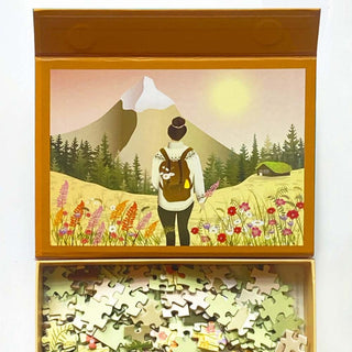 The ViSSEVASSE Hike with Me | 300 Piece Jigsaw Puzzle features an illustration of a woman with a backpack standing in a vibrant field of flowers, gazing toward mountains and a cabin, with puzzle pieces displayed beneath the image. This walking-themed puzzle comes in a sturdy box with magnetic closure for convenient storage and gifting.