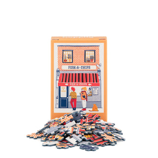 A box featuring an illustrated fish and chips shop from Happily Jigsaw Puzzles stands behind a pile of puzzle pieces, suggesting the pieces assemble into the picture on the box. The Fish & Chips | 99 Piece Mini Jigsaw Puzzle is a charming and delightful mini puzzle made from recycled board.