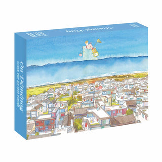 The WEVE Moving Day 1,000 Piece Jigsaw Puzzle features an illustrated cityscape with houses, mountains, and a trumpet-playing character floating above. Made with premium blue board for extra durability.