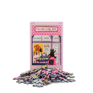 A Happily Jigsaw Puzzles box labeled “Feline Fine Art | 99 Piece Mini Jigsaw Puzzle” featuring cat-themed artwork stands behind a scattered pile of loose jigsaw pieces, inviting you to assemble this charming mini cat puzzle.