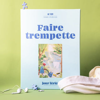 The Faire Trempette | 1,000 Piece Jigsaw Puzzle by Jour Férié features a riverside scene on a blue box, set against green with earphones, fabric, and puzzle pieces nearby. Made in France.
