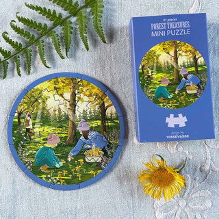 The Forest Treasure | 31 Piece Jigsaw Puzzle by ViSSEVASSE and its box feature an enchanting illustration of people gathering mushrooms in a sun-dappled forest. Nearby, a yellow flower and green fern rest on light-colored fabric, inviting you to creatively immerse yourself as you piece together this tranquil scene.