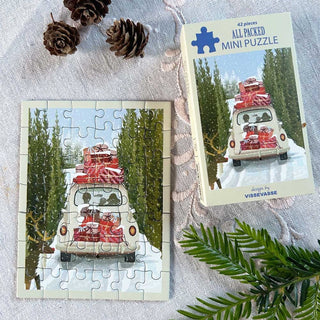 The All Packed | 42 Piece Jigsaw Puzzle by ViSSEVASSE, shown completed alongside its box, features a white car carrying red luggage as it drives through snowy pine trees. Pinecones and greenery are arranged nearby on a textured fabric surface.