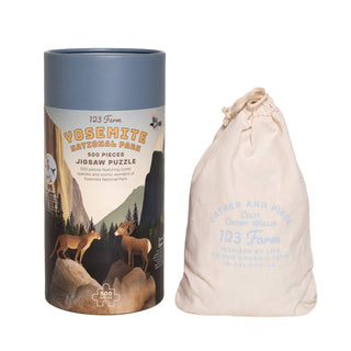 A cylindrical box labeled “Yosemite | 500 Piece Jigsaw Puzzle” by 123 Farm stands beside a blue-text drawstring bag, reflecting the splendor of this iconic location.