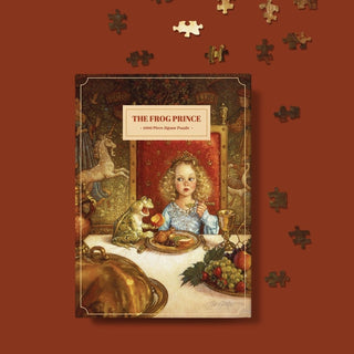 The Puzzle Theory’s The Frog Prince | 1,000 Piece Jigsaw Puzzle features Scott Gustafson’s illustration of a girl and frog at a lavish table with fruit and gold décor, plus loose pieces scattered nearby.