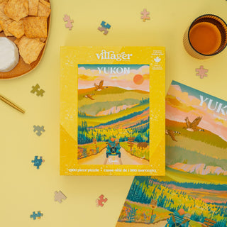 The Yukon Road Trip 1,000 Piece Jigsaw Puzzle by Villager Puzzles is shown on a yellow surface with scattered pieces, a drink, snacks, and a gold pen—an ideal Canada souvenir for puzzle lovers. Designed by Capri Sadler.